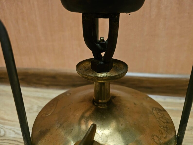 K&ouml;nnte beinhalten: Ein Vintage-Campingkocher aus Messing mit einem schwarzen Metallbrenner und einem schwarzen Metallst&auml;nder. Der Kocher hat einen runden Sockel und einen erh&ouml;hten Brenner.
