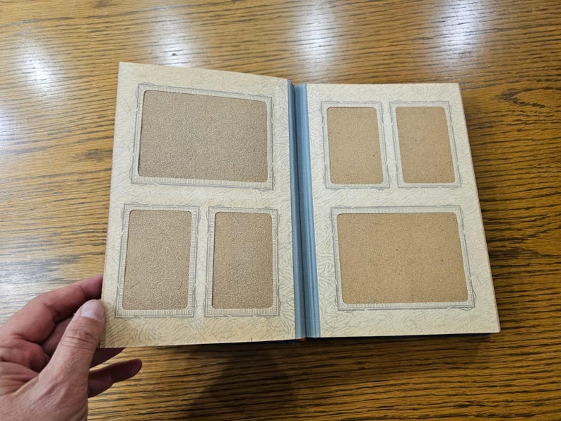 May include: A white photo album with a blue spine and five empty photo slots. The album has a decorative border around each photo slot.