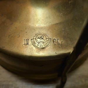K&ouml;nnte beinhalten: Nahaufnahme eines Campingkochers aus Messing mit einem Logo und den Zahlen "III" und "64", die auf den Boden gepr&auml;gt sind.