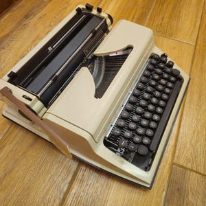 Może przedstawiać: Antyczna biała maszyna do pisania z czarną klawiaturą i dźwignią zwrotną karetki. Maszyna do pisania znajduje się na drewnianej powierzchni.