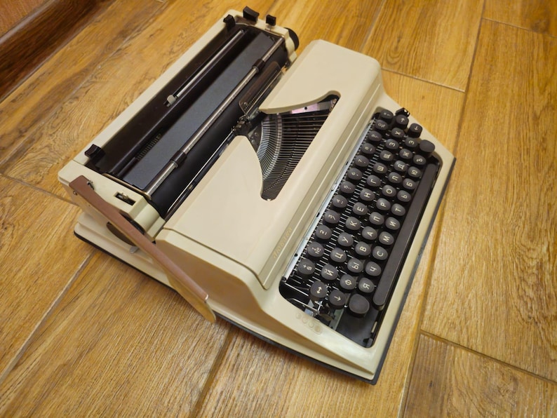 Może przedstawiać: Antyczna biała maszyna do pisania z czarną klawiaturą i dźwignią zwrotną karetki. Maszyna do pisania znajduje się na drewnianej powierzchni.