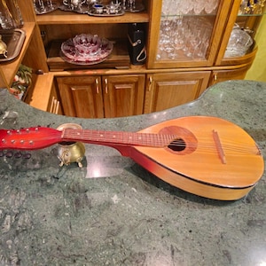 Puede incluir: Una mandolina de madera con clavijero rojo y cuerpo marrón claro descansa sobre una encimera de mármol verde. El instrumento tiene cuerdas y una boca. Una pequeña olla decorativa dorada está al lado de la mandolina.