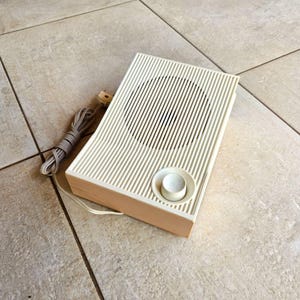 May include: A vintage white speaker with a round knob and a cord. The speaker has a wood trim around the edges.