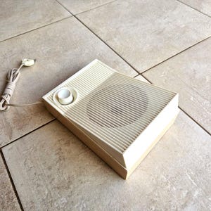 May include: A vintage, off-white, rectangular speaker with a round knob and a speaker grille. The speaker is sitting on a light brown tiled floor.