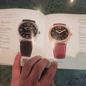 Pu&ograve; includere: Due orologi da polso con quadranti neri e indici orari in numeri romani. L'orologio a sinistra ha un cinturino in pelle nera e l'orologio a destra ha un cinturino in pelle marrone. Il testo "Radiomir" e "Radiomir ROSOVO ZOROTO" &egrave; visibile sulla pagina.