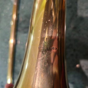 May include: Close-up of a gold trombone bell with the inscription "V 567 188* L 700" engraved on the side.