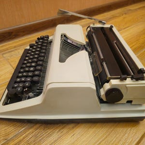 Może przedstawiać: Biała, zabytkowa maszyna do pisania z czarną klawiaturą i czarną dźwignią powrotu karetki. Maszyna do pisania stoi na drewnianej powierzchni.