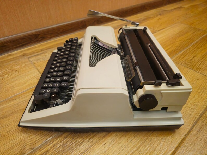 Może przedstawiać: Biała, zabytkowa maszyna do pisania z czarną klawiaturą i czarną dźwignią powrotu karetki. Maszyna do pisania stoi na drewnianej powierzchni.