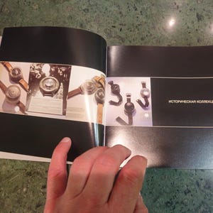 Pu&ograve; includere: Un libro aperto con una copertina nera. La pagina sinistra mostra un primo piano di diversi orologi vintage con cinturini in pelle. La pagina destra mostra un primo piano di diversi orologi neri con cinturini in metallo. Il testo "историческая коллекция" &egrave; stampato sulla pagina destra.