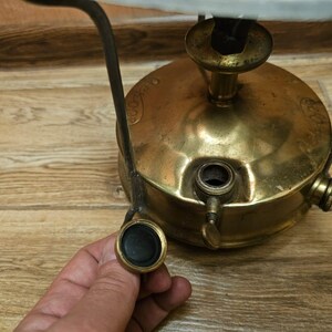 K&ouml;nnte beinhalten: Ein antiker Campingkocher aus Messing mit einem schwarzen Metallgriff und einem schwarzen Knopf. Der Kocher hat einen runden Sockel und einen zylindrischen oberen Teil mit einer kleinen &Ouml;ffnung f&uuml;r Brennstoff.