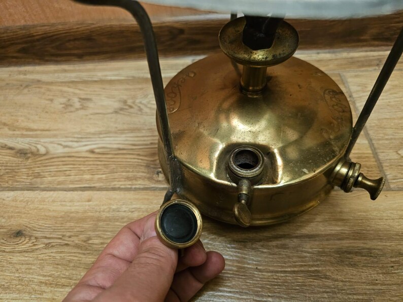 K&ouml;nnte beinhalten: Ein antiker Campingkocher aus Messing mit einem schwarzen Metallgriff und einem schwarzen Knopf. Der Kocher hat einen runden Sockel und einen zylindrischen oberen Teil mit einer kleinen &Ouml;ffnung f&uuml;r Brennstoff.