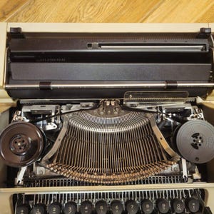 Może przedstawiać: Beige'owa zabytkowa maszyna do pisania z czarną szpulą taśmy i metalową listwą do pisania. Maszyna do pisania jest otwarta i gotowa do pisania.