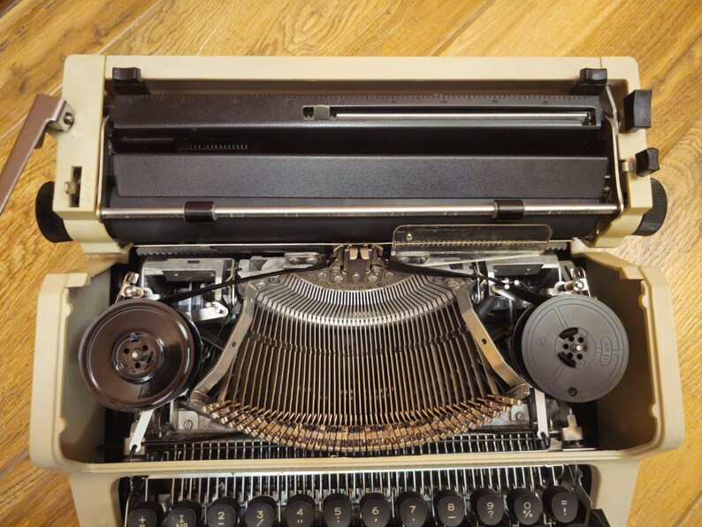 Może przedstawiać: Beige'owa zabytkowa maszyna do pisania z czarną szpulą taśmy i metalową listwą do pisania. Maszyna do pisania jest otwarta i gotowa do pisania.