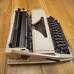 Może przedstawiać: Biała, zabytkowa maszyna do pisania z czarną klawiaturą i dźwignią zwrotki. Maszyna do pisania ma czarną metalową ramę i białe plastikowe obudowę.