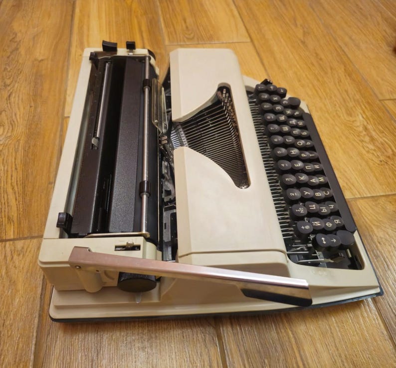 Może przedstawiać: Biała, zabytkowa maszyna do pisania z czarną klawiaturą i dźwignią zwrotki. Maszyna do pisania ma czarną metalową ramę i białe plastikowe obudowę.