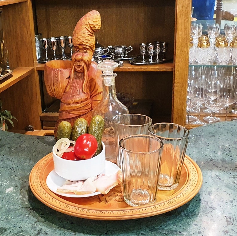 May include: A wooden carving of a man with a beard and a hat, holding a bowl. There are four glasses, a bottle, and a bowl of pickles and tomatoes on a wooden tray.