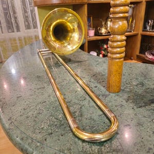 May include: A gold trombone with a bell-shaped mouthpiece rests on a green marble surface. The trombone is in a U-shape with the bell facing the camera.