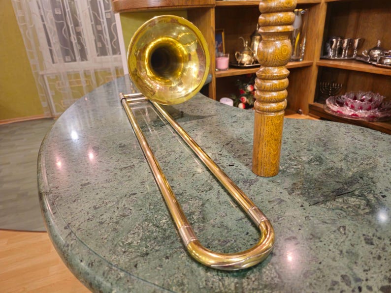 May include: A gold trombone with a bell-shaped mouthpiece rests on a green marble surface. The trombone is in a U-shape with the bell facing the camera.
