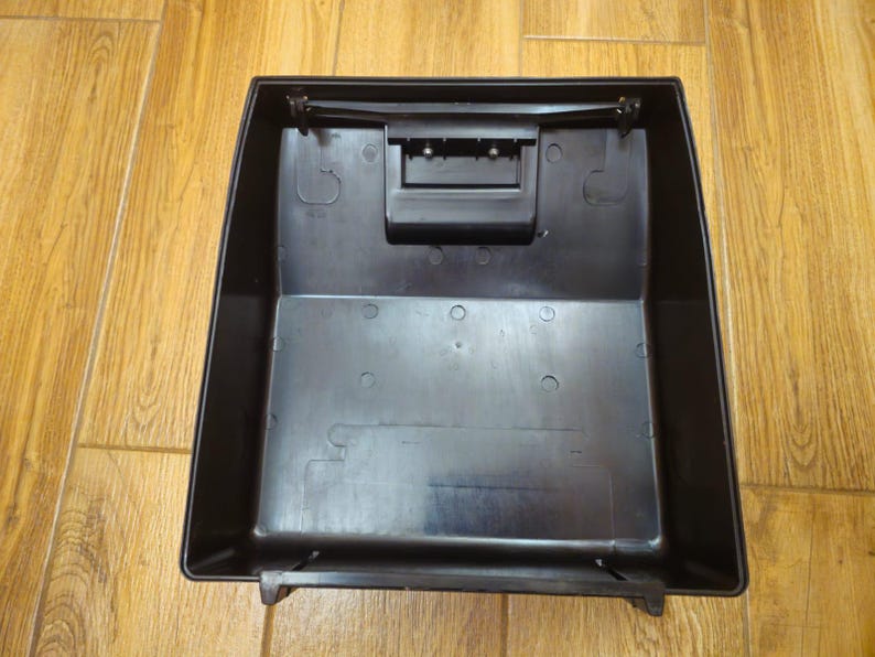 Może przedstawiać: Czarny plastikowy pojemnik do przechowywania o prostokątnym kształcie z pokrywką na zawiasach. Pojemnik ma mały prostokątny otw&oacute;r na g&oacute;rze.