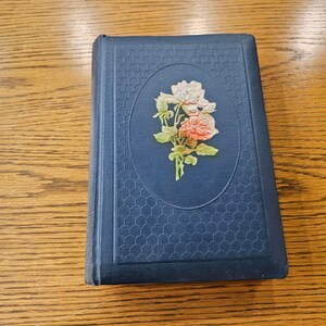 May include: A blue hardcover book with a floral design on the front cover. The design features a bouquet of pink and white roses with green leaves.
