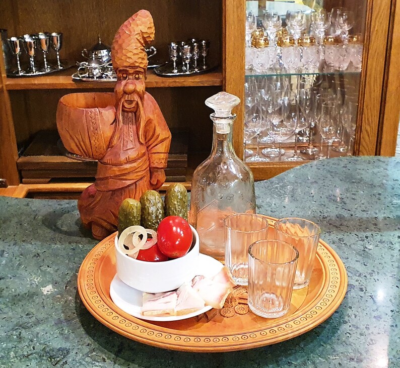 May include: A wooden carving of a man with a beard and a hat, holding a wooden bowl. The carving is on a wooden table with a glass bottle, glasses, pickles, and a bowl of food.