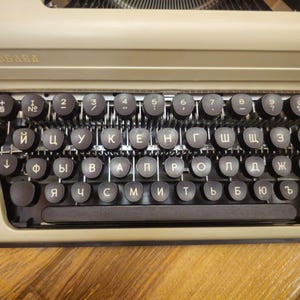 Może przedstawiać: Antyczna maszyna do pisania z beżowym korpusem i czarnymi klawiszami. Klawisze są oznaczone literami cyrylicy. Maszyna do pisania ma nazwę marki "ЛЮБАВА" na g&oacute;rze.