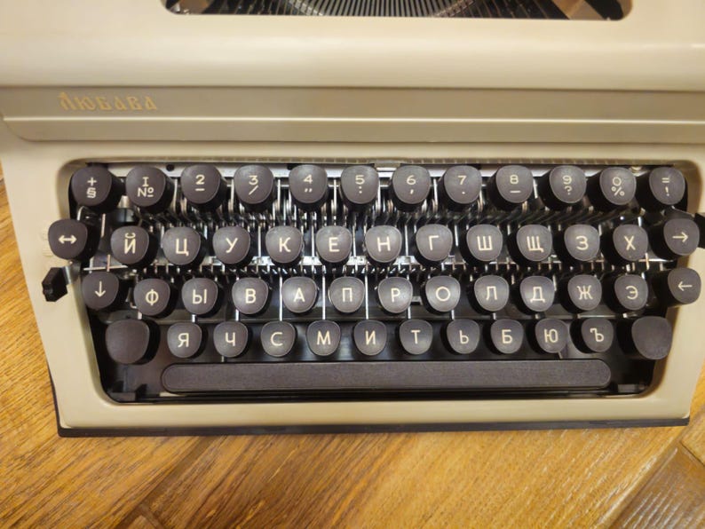 Może przedstawiać: Antyczna maszyna do pisania z beżowym korpusem i czarnymi klawiszami. Klawisze są oznaczone literami cyrylicy. Maszyna do pisania ma nazwę marki "ЛЮБАВА" na g&oacute;rze.