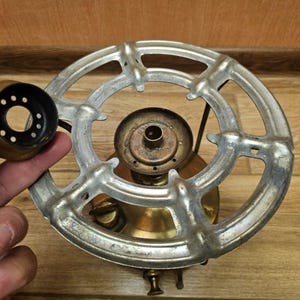 K&ouml;nnte beinhalten: Ein antiker Herdbrenner aus Messing mit einem runden Metallrost und einer zentralen Brenner&ouml;ffnung. Der Brenner hat ein dekoratives Design mit einer Reihe konzentrischer Kreise und einem erh&ouml;hten Rand.
