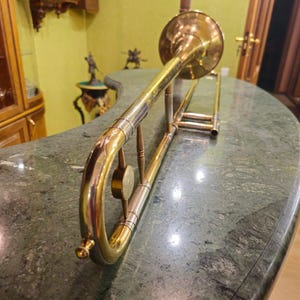 May include: A gold-colored trombone with a bell-shaped mouthpiece and a slide. The trombone is lying on a green marble surface.