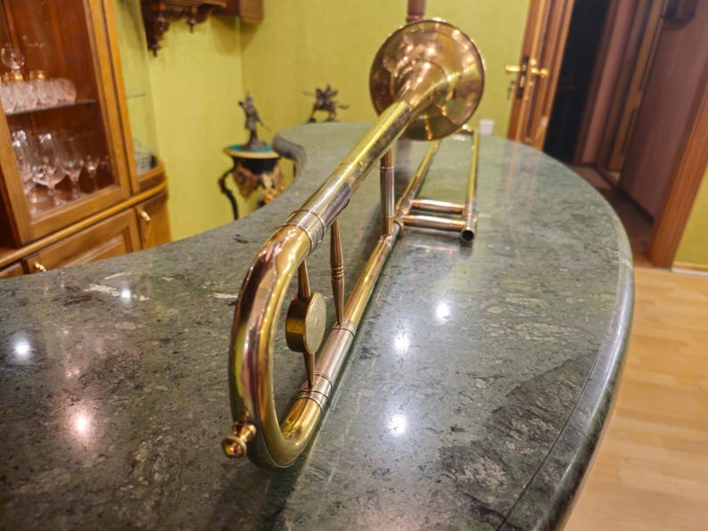 May include: A gold-colored trombone with a bell-shaped mouthpiece and a slide. The trombone is lying on a green marble surface.