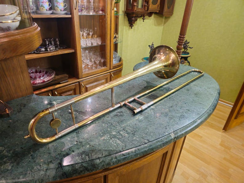 May include: A gold-colored trombone with a silver slide. The trombone is lying on a green marble surface.