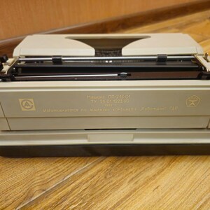 Może przedstawiać: Beżowa maszyna do pisania z czarną dźwignią powrotu karetki i czarną i srebrną klawiaturą. Maszyna do pisania ma etykietę z napisem "Машина ПП-215-01 ТУ 25-01-1223-82 Изготовляется по лицензии "Kenbi" Роботрон ГДР".