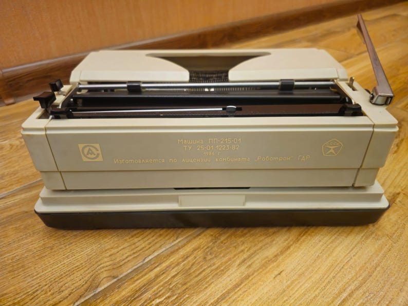 Może przedstawiać: Beżowa maszyna do pisania z czarną dźwignią powrotu karetki i czarną i srebrną klawiaturą. Maszyna do pisania ma etykietę z napisem "Машина ПП-215-01 ТУ 25-01-1223-82 Изготовляется по лицензии "Kenbi" Роботрон ГДР".