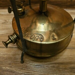 K&ouml;nnte beinhalten: Campingkocher aus Messing mit einem schwarzen Metallst&auml;nder und einem Etikett mit der Aufschrift "Primus 1".