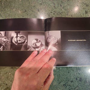 Pu&ograve; includere: Un libro aperto con pagine nere e sfondo bianco. Il libro &egrave; aperto a una pagina con tre immagini di parti di orologio in lavorazione. Il testo "kollektion - manufaktur" &egrave; stampato sulla pagina destra.