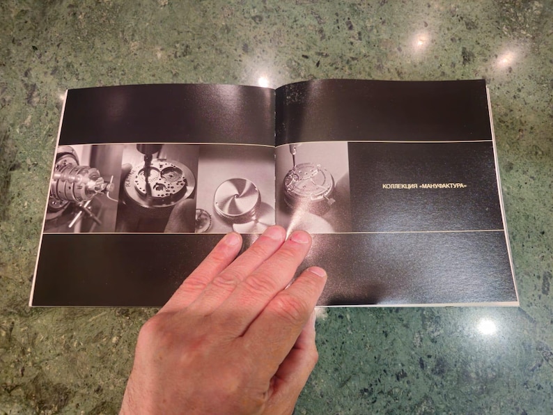 Pu&ograve; includere: Un libro aperto con pagine nere e sfondo bianco. Il libro &egrave; aperto a una pagina con tre immagini di parti di orologio in lavorazione. Il testo "kollektion - manufaktur" &egrave; stampato sulla pagina destra.