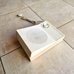 May include: A vintage white and beige speaker with a round knob on the top. The speaker has a grill covering the speaker cone.