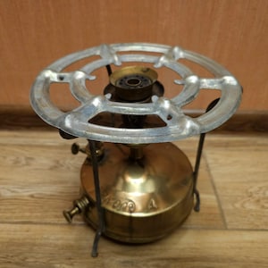 K&ouml;nnte beinhalten: Ein antiker Campingkocher aus Messing mit einem Metallrost und drei Beinen. Der Kocher hat einen runden Sockel und einen runden Brenner mit einem kleinen Loch in der Mitte.