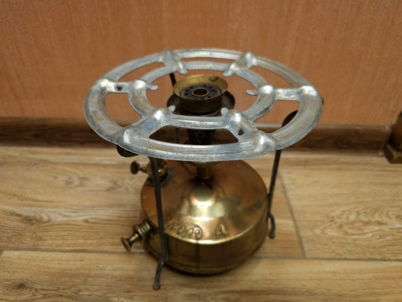 K&ouml;nnte beinhalten: Ein antiker Campingkocher aus Messing mit einem Metallrost und drei Beinen. Der Kocher hat einen runden Sockel und einen runden Brenner mit einem kleinen Loch in der Mitte.