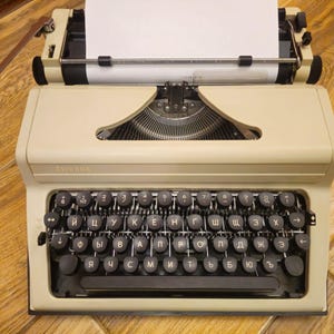 Może przedstawiać: Antyczna beżowa maszyna do pisania z czarną klawiaturą i białą kartką papieru w karetce. Klawiatura ma cyrylicę.