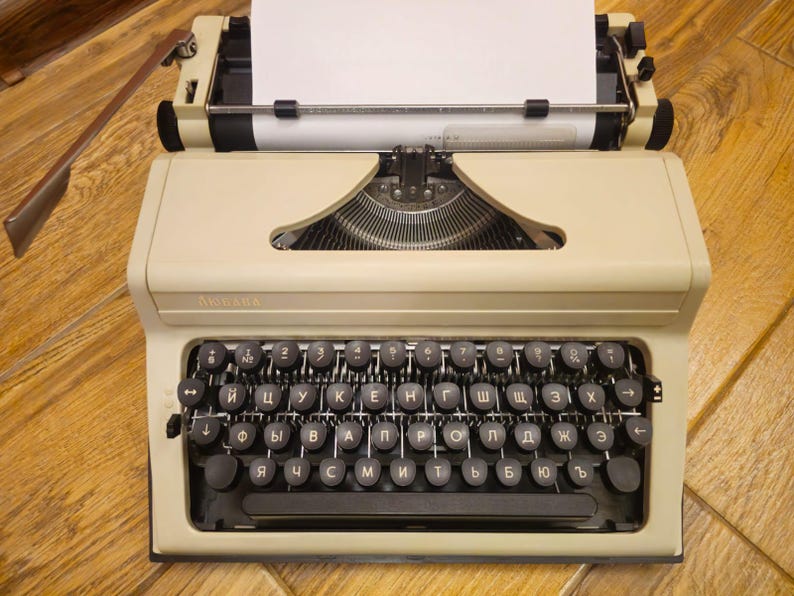 Może przedstawiać: Antyczna beżowa maszyna do pisania z czarną klawiaturą i białą kartką papieru w karetce. Klawiatura ma cyrylicę.