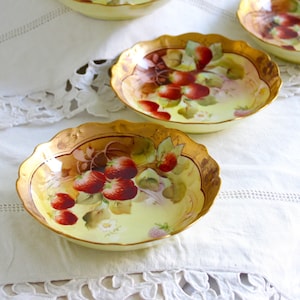 May include: Five antique porcelain bowls with gold trim and a hand-painted design of strawberries and leaves. The bowls are on a white tablecloth with lace trim.