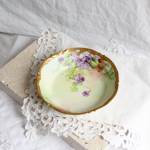 May include: A vintage porcelain bowl with a delicate floral design. The bowl features a pale green background with purple flowers and gold accents around the rim.
