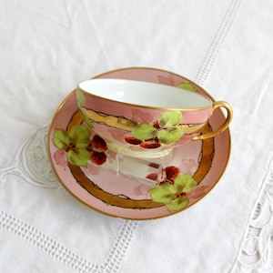 May include: A pink teacup and saucer with a gold rim and painted floral design. The floral design features green leaves and red berries.