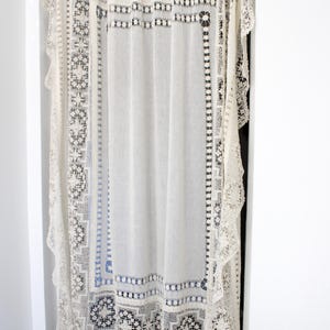May include: An off-white lace curtain with detailed geometric patterns and scalloped edges. The curtain hangs in a doorway, with a matching valance at the top. The delicate design adds a vintage touch to the room.