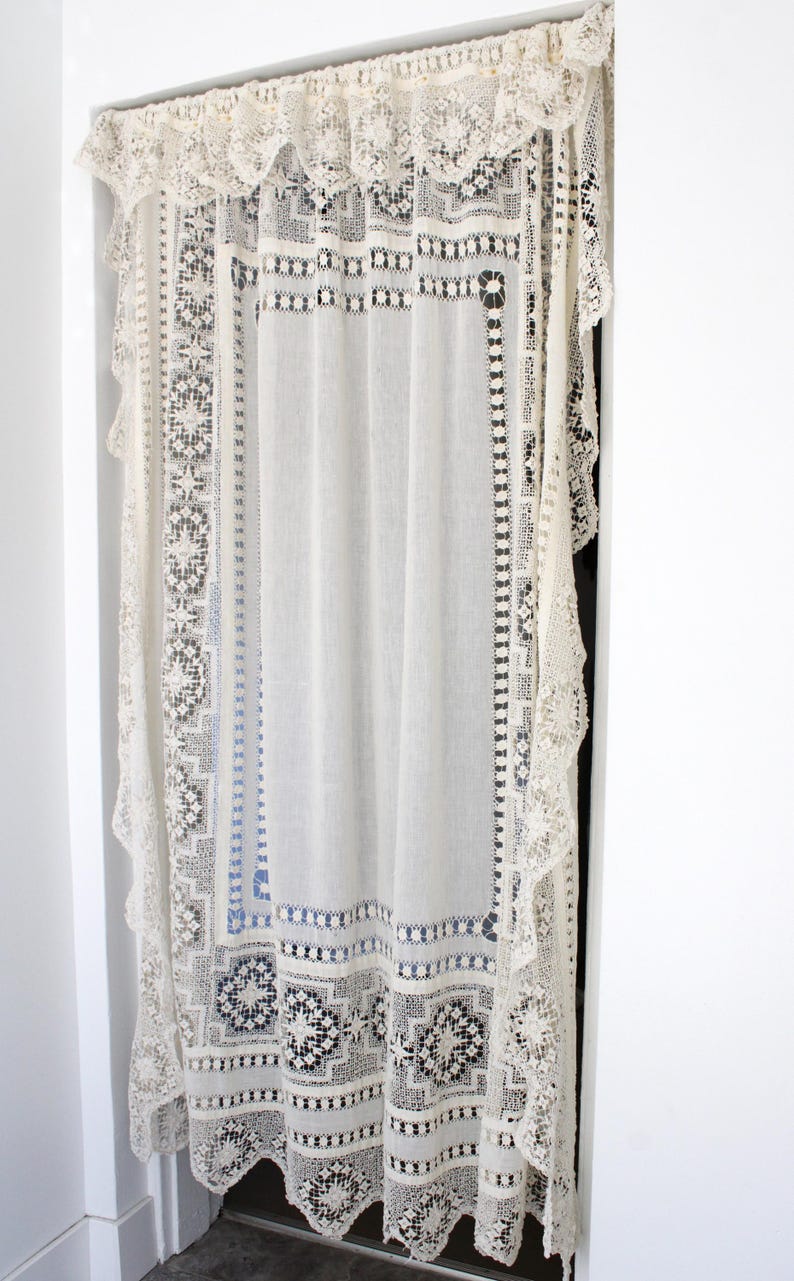 May include: An off-white lace curtain with detailed geometric patterns and scalloped edges. The curtain hangs in a doorway, with a matching valance at the top. The delicate design adds a vintage touch to the room.