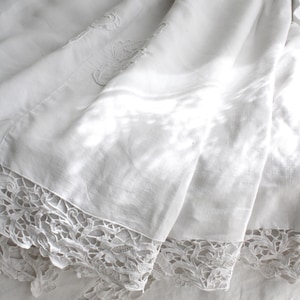 May include: White linen bedding with detailed lace edging. The fabric is folded, highlighting the texture and design of the lace. The bedding looks soft and delicate, with gentle shadows.