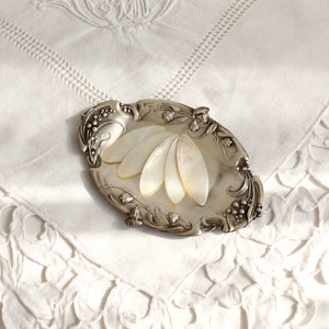 May include: A silver-toned decorative dish with a mother-of-pearl inlay in the shape of a fan. The dish is ornate with floral details and is sitting on a white lace tablecloth.