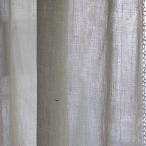 May include: Close-up of sheer, off-white curtains. The fabric has a fine, open weave, allowing light to filter through. The curtains are slightly textured, with subtle variations in tone. The edges of the curtains are softly draped, creating a gentle, flowing effect.