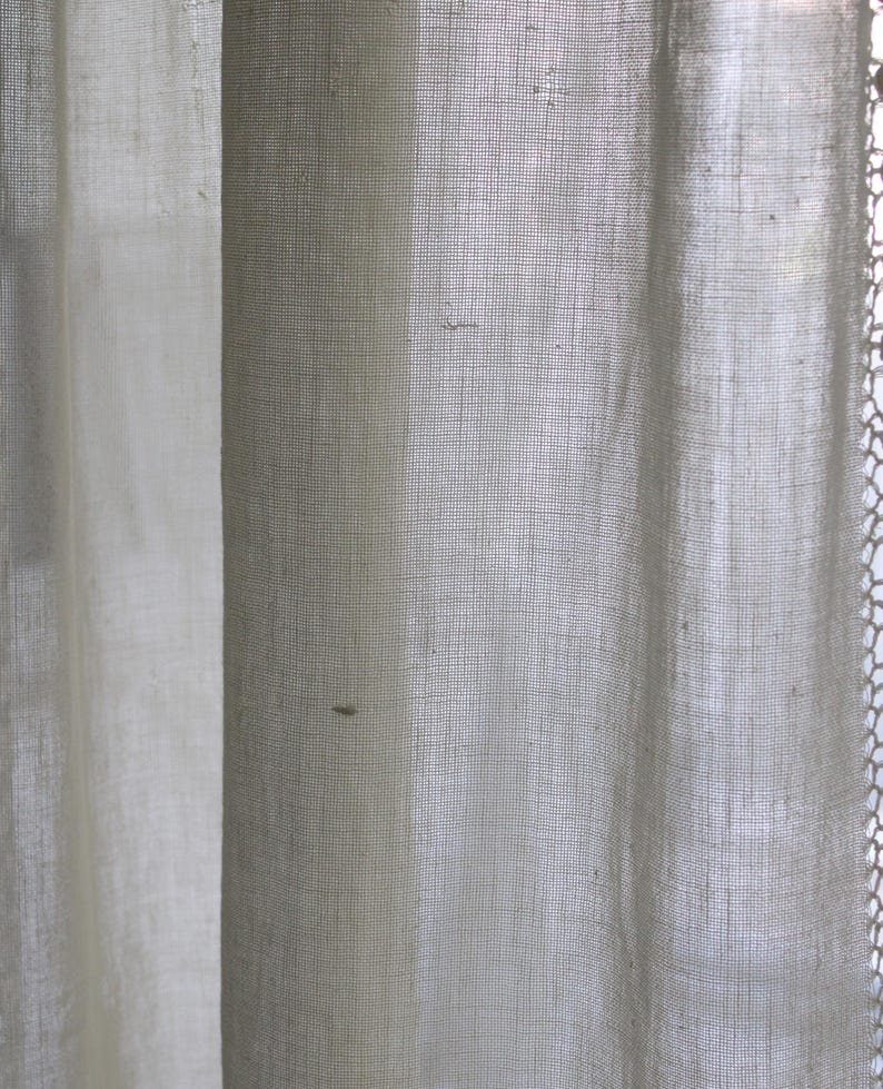 May include: Close-up of sheer, off-white curtains. The fabric has a fine, open weave, allowing light to filter through. The curtains are slightly textured, with subtle variations in tone. The edges of the curtains are softly draped, creating a gentle, flowing effect.